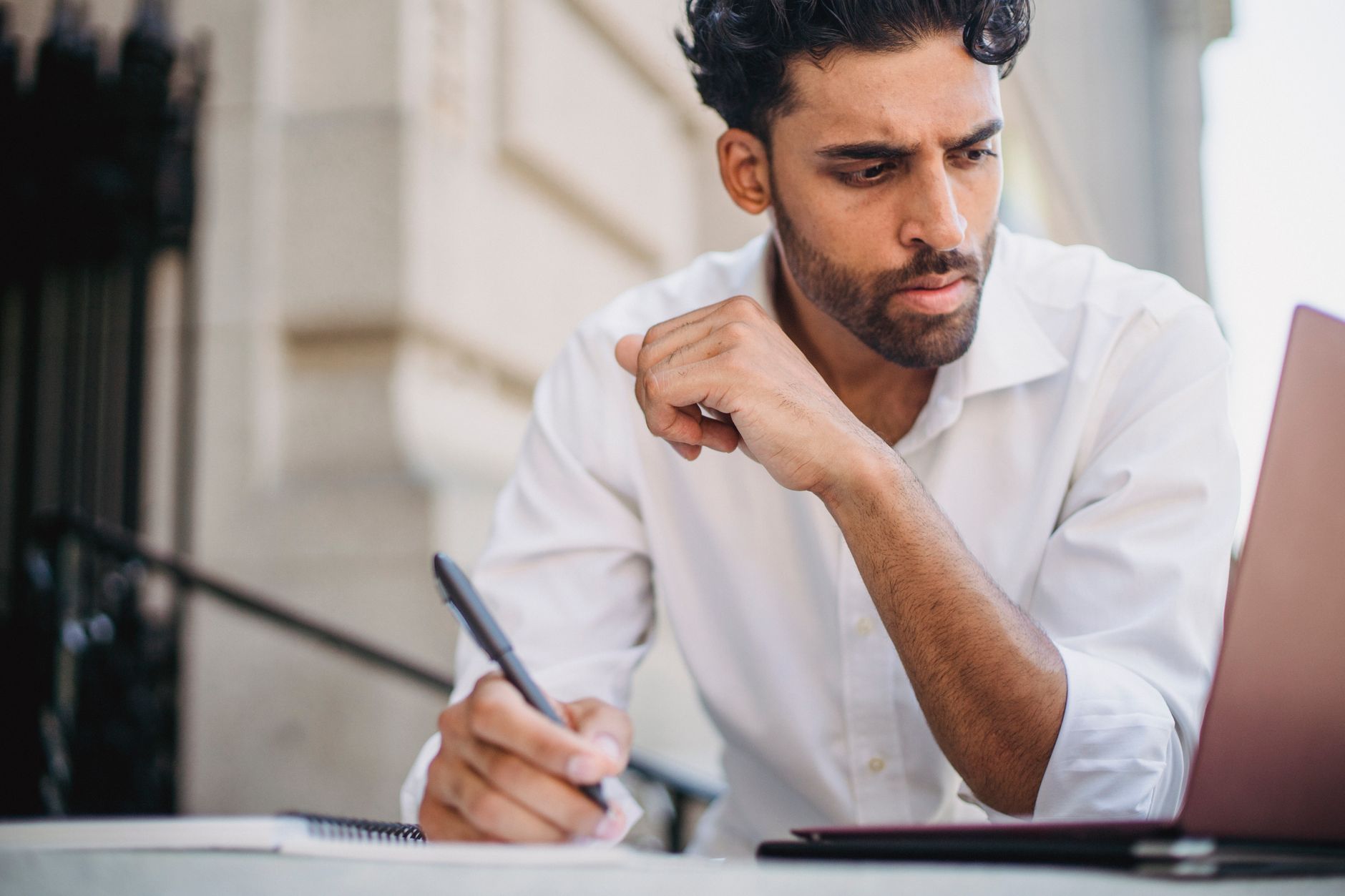 A person talks on the phone while taking notes at a desk with a serious expression.
